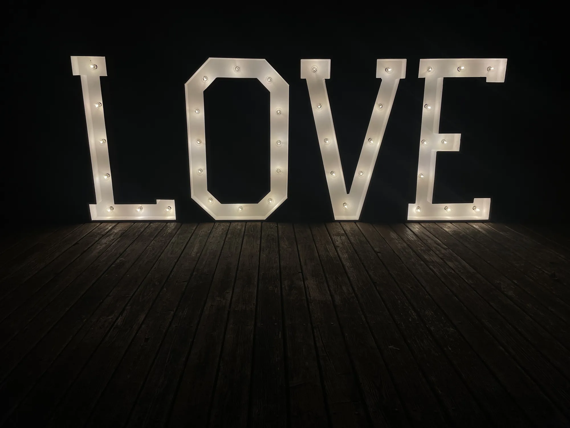 Four white marquee letters spelling LOVE, lit with warm bulbs on a wooden deck at night